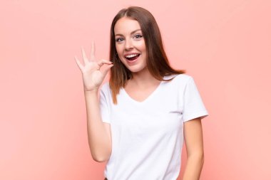 Genç kızıl saçlı kadın kendini başarılı ve tatmin olmuş hissediyor, ağzı açık gülümsüyor, düz duvara el işareti yapıyor.