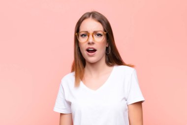 Genç kızıl saçlı kadın dehşete kapılmış ve şok olmuş hissediyor, ağzı sonuna kadar açık, düz duvara karşı şaşkınlık içinde.