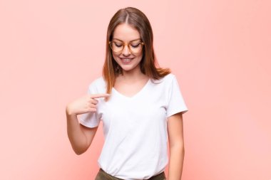 Genç kızıl saçlı kadın neşeyle ve gelişigüzel gülümsüyor, aşağıya bakıyor ve düz duvara doğru göğsünü gösteriyor.