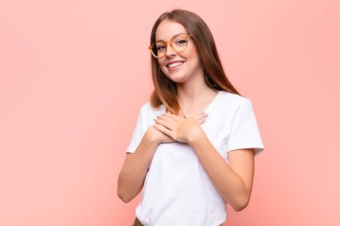 Genç kızıl saçlı kadın romantik, mutlu ve aşık, neşeyle gülümsüyor ve düz duvara karşı kalbe yakın el ele tutuşuyor.