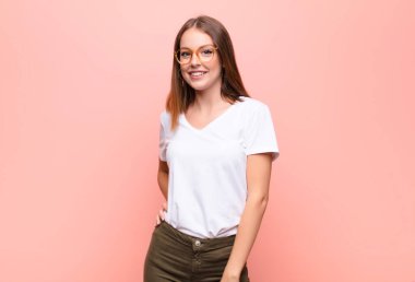 Genç kızıl saçlı kadın neşeli bir şekilde gülümsüyor. Olumlu, mutlu, kendinden emin ve gevşek bir ifadeyle düz duvara yaslanıyor.