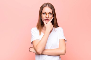 Genç kızıl saçlı kadın ciddi, düşünceli ve güvensiz görünüyor, bir kolu çapraz ve çene üzerinde, düz duvara karşı seçenekleri tartıyor