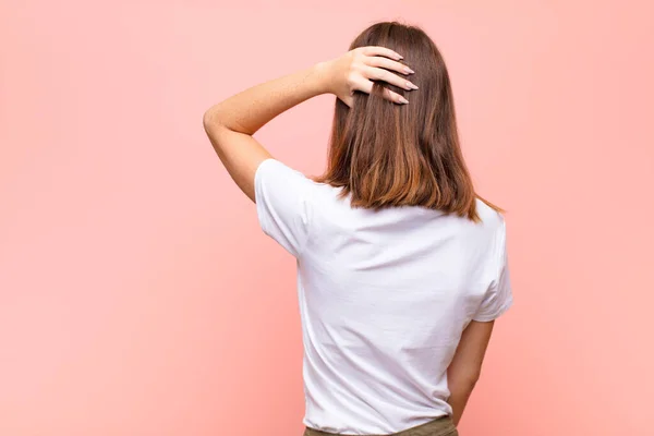 young red head woman thinking or doubting, scratching head, feeling puzzled and confused, back or rear view against flat wall