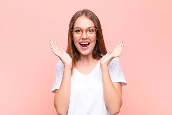 young red head woman feeling shocked and excited, laughing, amazed and happy because of an unexpected surprise against flat wall