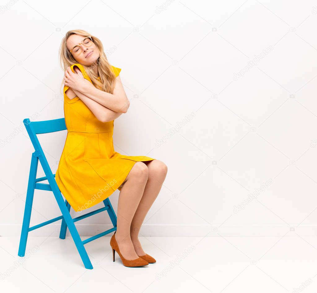 young pretty blonde woman feeling in love, smiling, cuddling and hugging self, staying single, being selfish and egocentric against flat color wall