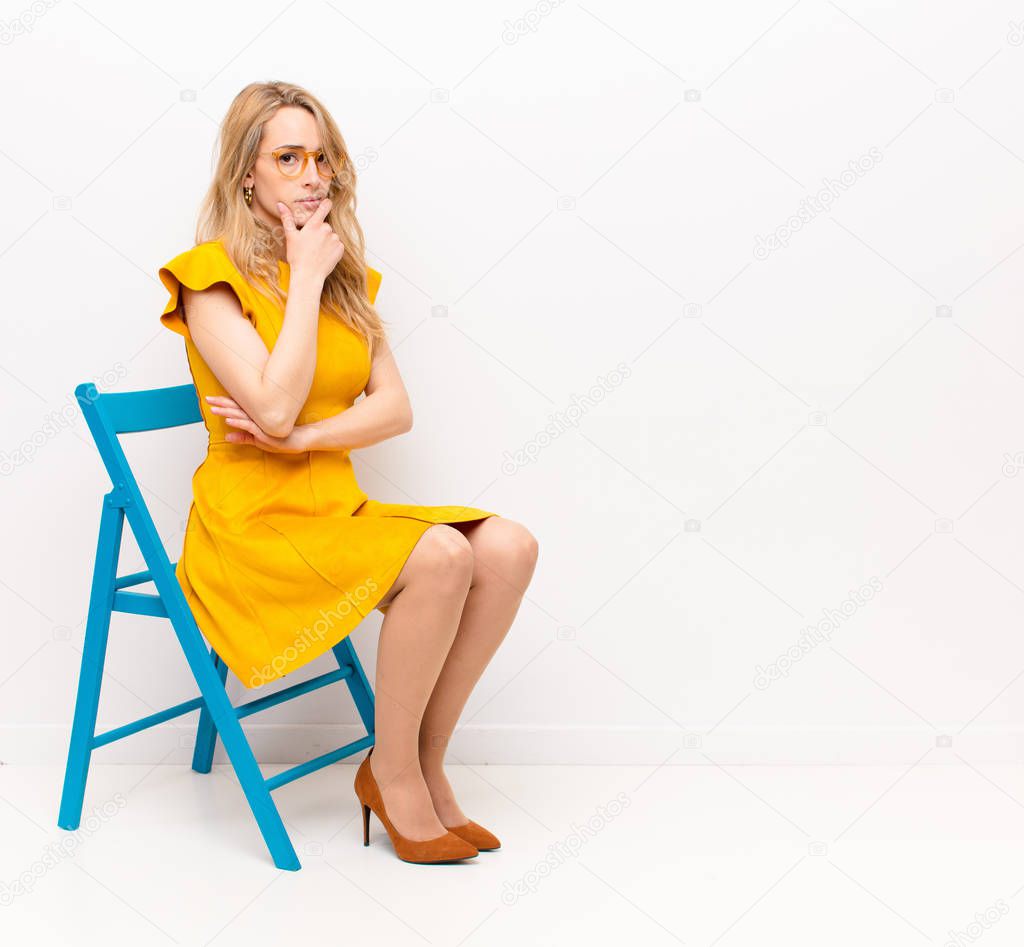 young pretty blonde woman looking serious, thoughtful and distrustful, with one arm crossed and hand on chin, weighting options against flat color wall