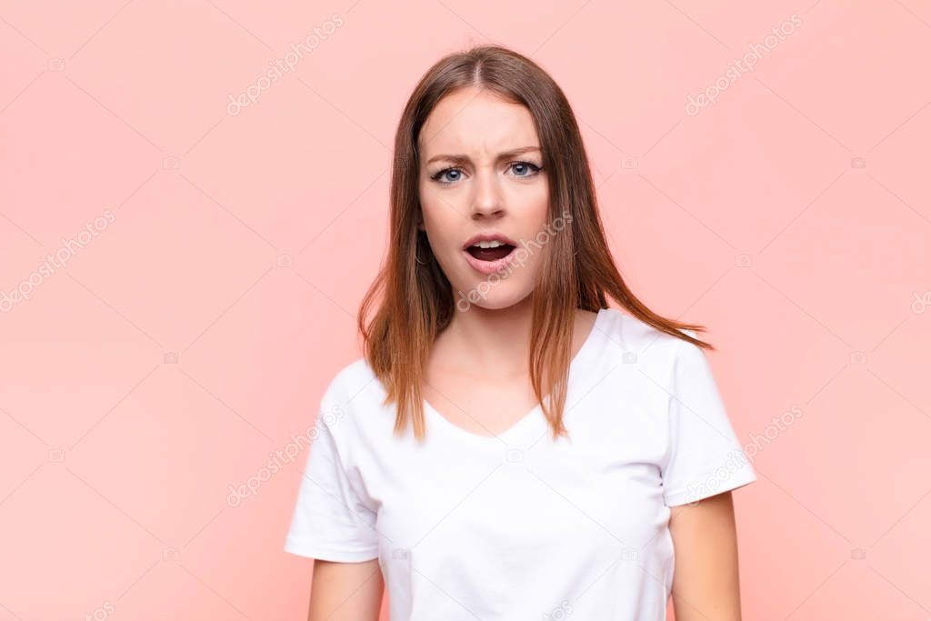 young red head woman looking shocked, angry, annoyed or disappointed, open mouthed and furious against flat wall