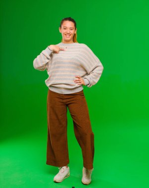 Genç ve güzel Latin kadın mutlu, gururlu ve şaşırmış görünüyor, neşeli bir şekilde kendini gösteriyor, kendinden emin ve yeşil duvara karşı yüce hissediyor.