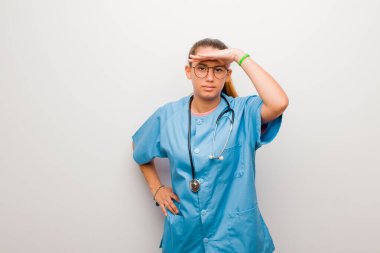 Genç Latin hemşire şaşkınlık ve şaşkınlık içinde bakıyor, el üstünde uzak bakıyor, beyaz duvarın karşısında izliyor ya da arıyor