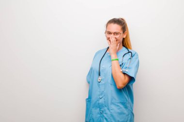 Genç Latin hemşire mutlu, neşeli, şanslı ve şaşırmış görünüyor. İki elini de beyaz duvara dayayıp ağzını kapatmış.