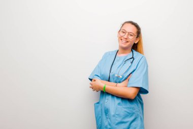 Genç Latin hemşire kollarını kavuşturup mutlu bir şekilde gülüyor. Beyaz duvara karşı rahat, olumlu ve tatmin edici bir pozla.