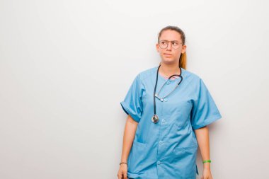 Genç Latin hemşire şaşkın ve şaşkın görünüyor, bir sorunu çözmeye çalışıyor ya da beyaz duvara karşı düşünüyor.