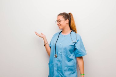 Genç Latin hemşire kendini mutlu hissediyor ve gülümsüyor. Beyaz duvara karşı elinde tuttuğu bir nesneye ya da konsepte bakıyor.