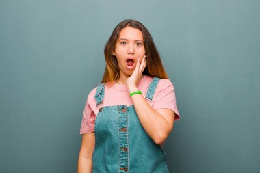 Genç ve güzel Latin bir kadın şok ve inançsızlık içinde, elleri yanak ve kolları çapraz, sersemlemiş ve şaşkınlık içinde grunge duvarına karşı