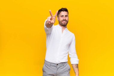 Genç adam gülümsüyor ve mutlu görünüyor, kaygısız ve pozitif, bir eliyle düz bir duvara karşı zafer ya da barış işareti yapıyor.