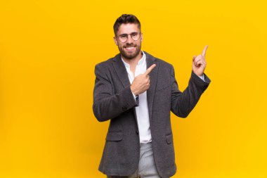 Genç adam mutlu bir şekilde gülümsüyor ve iki eliyle yukarı ve yana doğru işaret ediyor fotokopi uzayında düz renkli duvara karşı nesne gösteriyor.