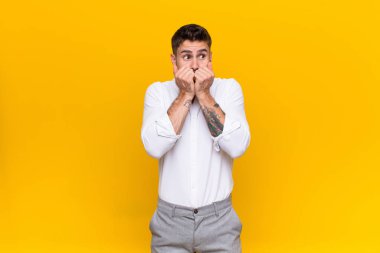 Genç adam endişeli, gergin, stresli ve korkmuş görünüyor, tırnak yiyor ve düz renkli duvara karşı yanal kopyalama alanı arıyor.