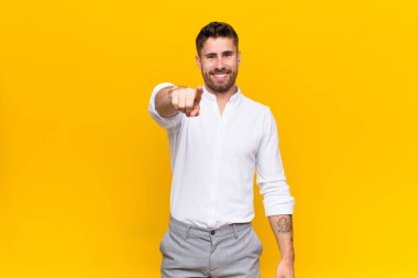 Genç adam kameraya tatmin olmuş, kendine güvenen, dost canlısı bir gülümsemeyle bakıyor, düz renkli duvara karşı seni seçiyor.