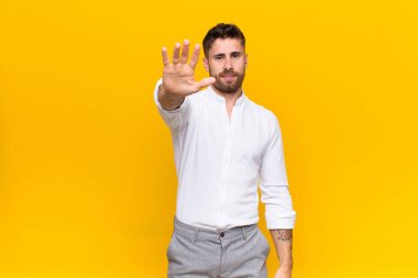 Genç adam ciddi, sert, hoşnutsuz ve kızgın görünüyor. Avuç içi açık, düz renkli duvara karşı jest yapıyor.