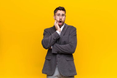 Genç adam şok ve inançsızlık içinde ağzı açık, elleri yanak ve kolları çapraz, sersemlemiş ve düz renkli duvara karşı şaşkın ve şaşkın hissediyor
