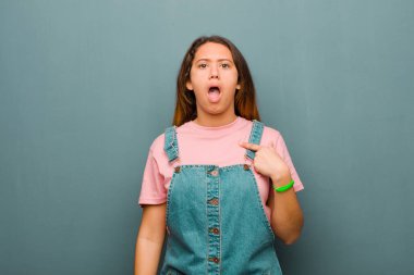 Genç ve güzel Latin kadın şok olmuş ve şaşırmış görünüyor ağzı sonuna kadar açık, grunge duvarına karşı kendini işaret ederek