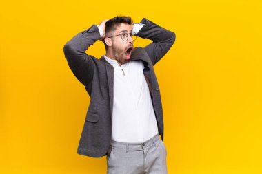 Ağzı açık genç bir adam, korkunç bir hata yüzünden dehşete düşmüş ve şok olmuş görünüyor, düz renkli duvara karşı el kaldırıyor.