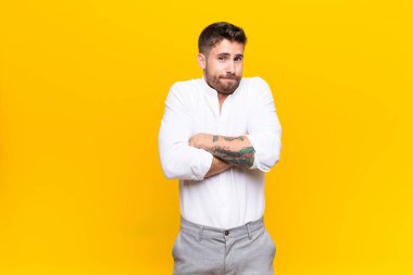 Genç adam omuz silkiyor, kafası karışık ve kararsız, kolları çapraz ve şaşkın bir şekilde düz duvara bakıyor.