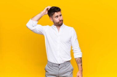 Genç adam şaşkın ve şaşkın hissediyor, başını kaşıyor ve düz renkli duvara bakıyor.