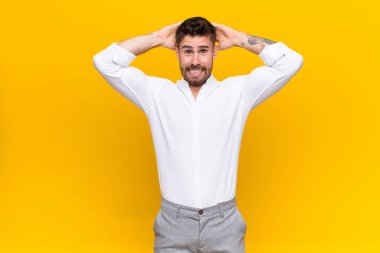 Genç adam kendini stresli, endişeli, endişeli ya da korkmuş hissediyor, elleri başının üstünde, düz renkli duvara karşı panikliyor.