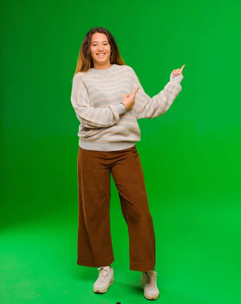 young pretty latin woman smiling happily and pointing to side and upwards with both hands showing object in copy space against green wall
