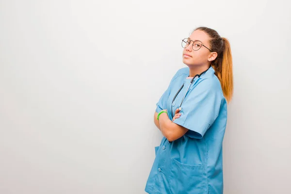 young latin nurse shrugging, feeling confused and uncertain, doubting with arms crossed and puzzled look against white wall