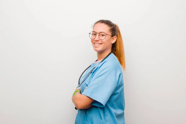 young latin nurse smiling to camera with crossed arms and a happy, confident, satisfied expression, lateral view against white wall