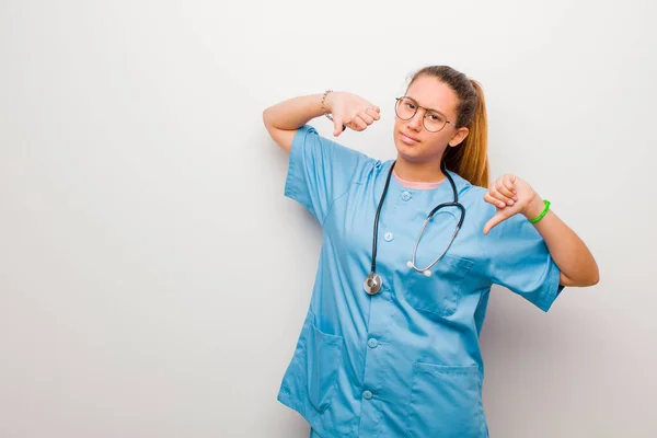 young latin nurse looking sad, disappointed or angry, showing thumbs down in disagreement, feeling frustrated against white wall