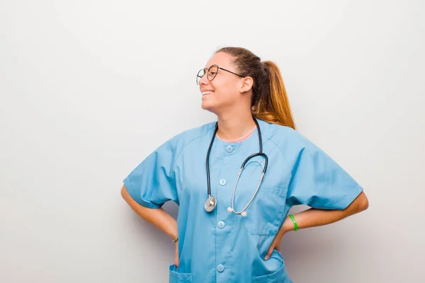 young latin nurse looking happy, cheerful and confident, smiling proudly and looking to side with both hands on hips against white wall