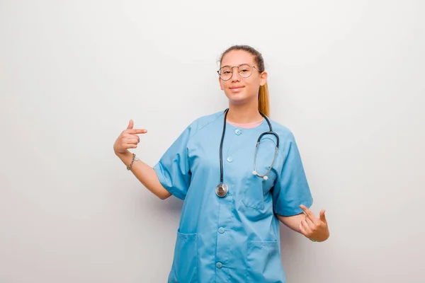 young latin nurse looking proud, arrogant, happy, surprised and satisfied, pointing to self, feeling like a winner against white wall