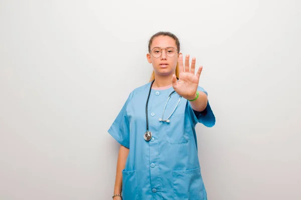 young latin nurse looking serious, stern, displeased and angry showing open palm making stop gesture against white wall