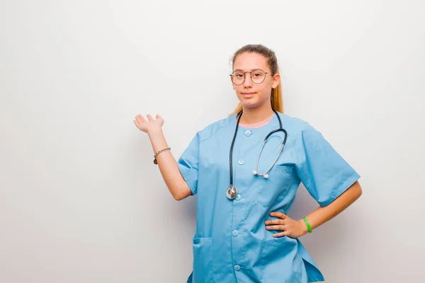 young latin nurse smiling, feeling confident, successful and happy, showing concept or idea on copy space on the side against white wall