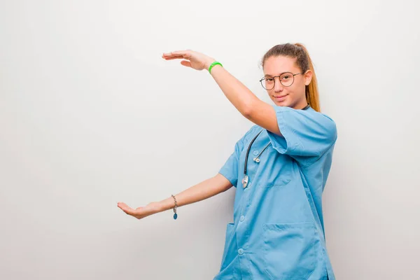 young latin nurse smiling, feeling happy, positive and satisfied, holding or showing object or concept on copy space against white wall