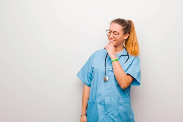 young latin nurse smiling with a happy, confident expression with hand on chin, wondering and looking to the side against white wall