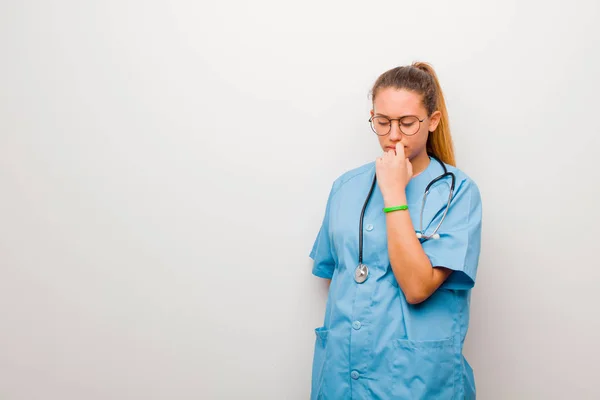 young latin nurse feeling serious, thoughtful and concerned, staring sideways with hand pressed against chin against white wall