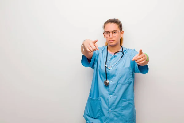 young latin nurse pointing forward at camera with both fingers and angry expression, telling you to do your duty against white wall
