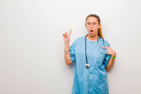 young latin nurse feeling proud and surprised, pointing to self confidently, feeling like successful number one against white wall