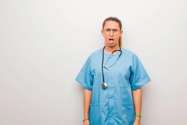 young latin nurse feeling terrified and shocked, with mouth wide open in surprise against white wall