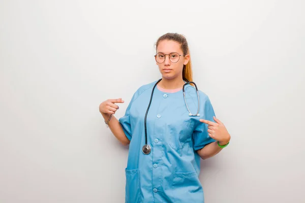 若いラテン語の看護師は誇りに思っています 白壁に対して両手で胸を正とカジュアルな指す — ストック写真