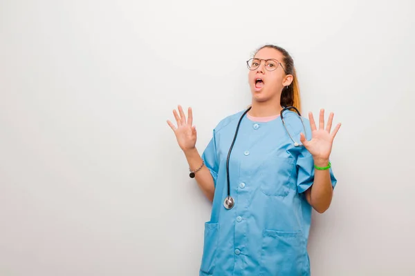 young latin nurse feeling stupefied and scared, fearing something frightening, with hands open up front saying stay away against white wall