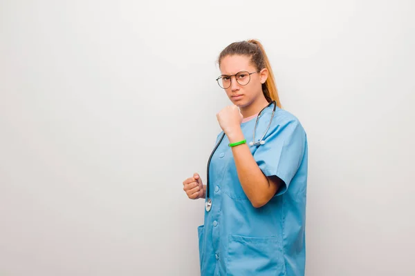 young latin nurse looking confident, angry, strong and aggressive, with fists ready to fight in boxing position against white wall