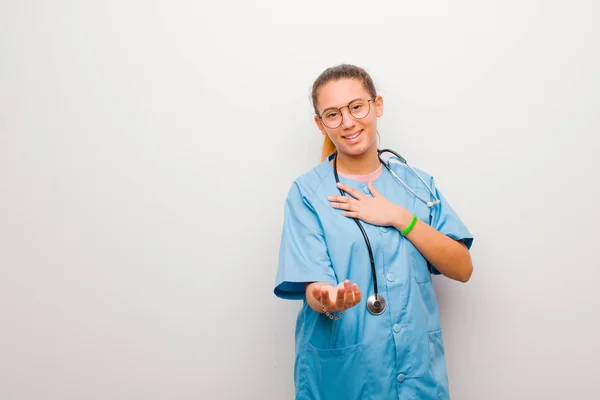 young latin nurse feeling happy and in love, smiling with one hand next to heart and the other stretched up front against white wall