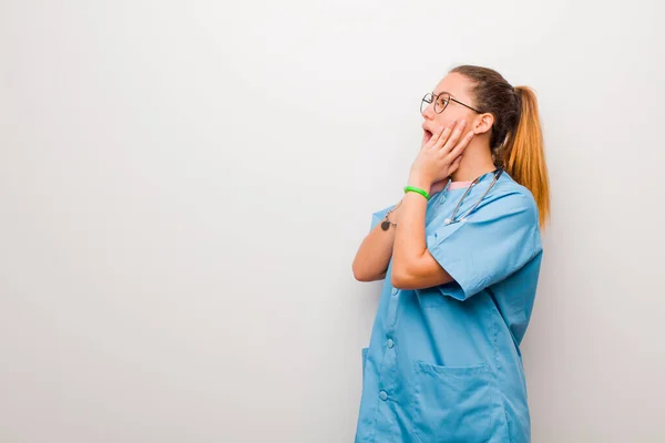 young latin nurse feeling happy, excited and surprised, looking to the side with both hands on face against white wall