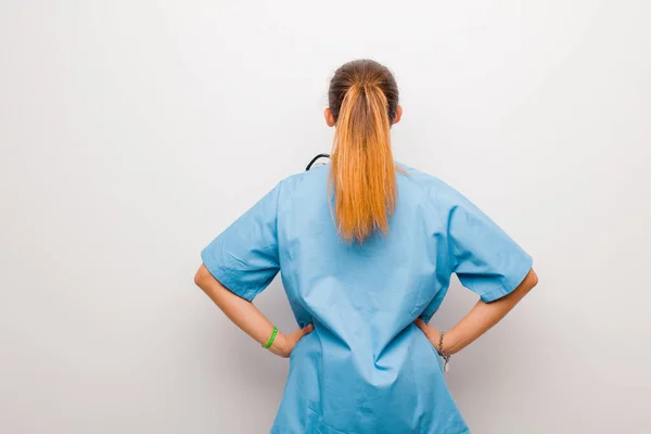 young latin nurse feeling confused or full or doubts and questions, wondering, with hands on hips, rear view against white wall