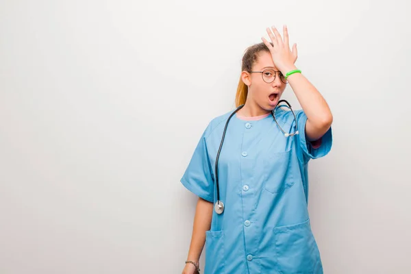 young latin nurse raising palm to forehead thinking oops, after making a stupid mistake or remembering, feeling dumb against white wall